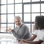 cheerful senior gray haired mentor supporting young black colleague in office