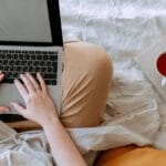 crop person using laptop with empty screen in bed near cup of tea placed on notebook