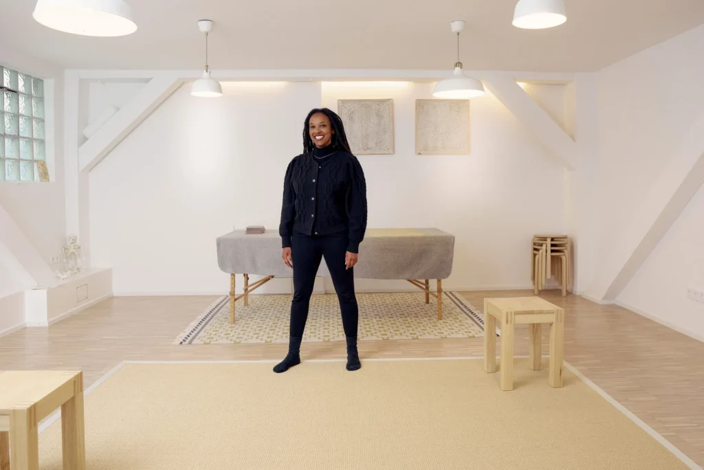 Eine Frau sitzt entspannt auf einem Stuhl und lehnt sich leicht zur Seite. Sie hat lange, dunkle Dreadlocks und trägt ein schwarzes Outfit. Ihre Hände sind locker ineinander verschränkt, und sie schaut mit einem ruhigen, freundlichen Ausdruck in die Kamera. Der Hintergrund ist hell und schlicht, mit ein paar gestapelten Hockern im Raum.