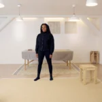 Eine Frau sitzt entspannt auf einem Stuhl und lehnt sich leicht zur Seite. Sie hat lange, dunkle Dreadlocks und trägt ein schwarzes Outfit. Ihre Hände sind locker ineinander verschränkt, und sie schaut mit einem ruhigen, freundlichen Ausdruck in die Kamera. Der Hintergrund ist hell und schlicht, mit ein paar gestapelten Hockern im Raum.