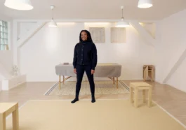Eine Frau sitzt entspannt auf einem Stuhl und lehnt sich leicht zur Seite. Sie hat lange, dunkle Dreadlocks und trägt ein schwarzes Outfit. Ihre Hände sind locker ineinander verschränkt, und sie schaut mit einem ruhigen, freundlichen Ausdruck in die Kamera. Der Hintergrund ist hell und schlicht, mit ein paar gestapelten Hockern im Raum.