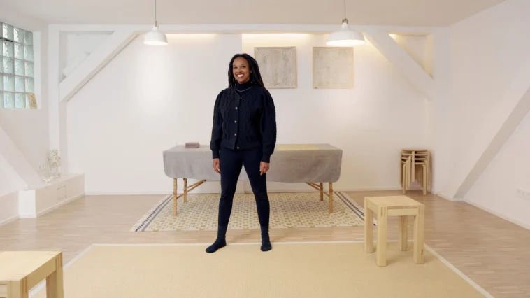 Eine Frau sitzt entspannt auf einem Stuhl und lehnt sich leicht zur Seite. Sie hat lange, dunkle Dreadlocks und trägt ein schwarzes Outfit. Ihre Hände sind locker ineinander verschränkt, und sie schaut mit einem ruhigen, freundlichen Ausdruck in die Kamera. Der Hintergrund ist hell und schlicht, mit ein paar gestapelten Hockern im Raum.