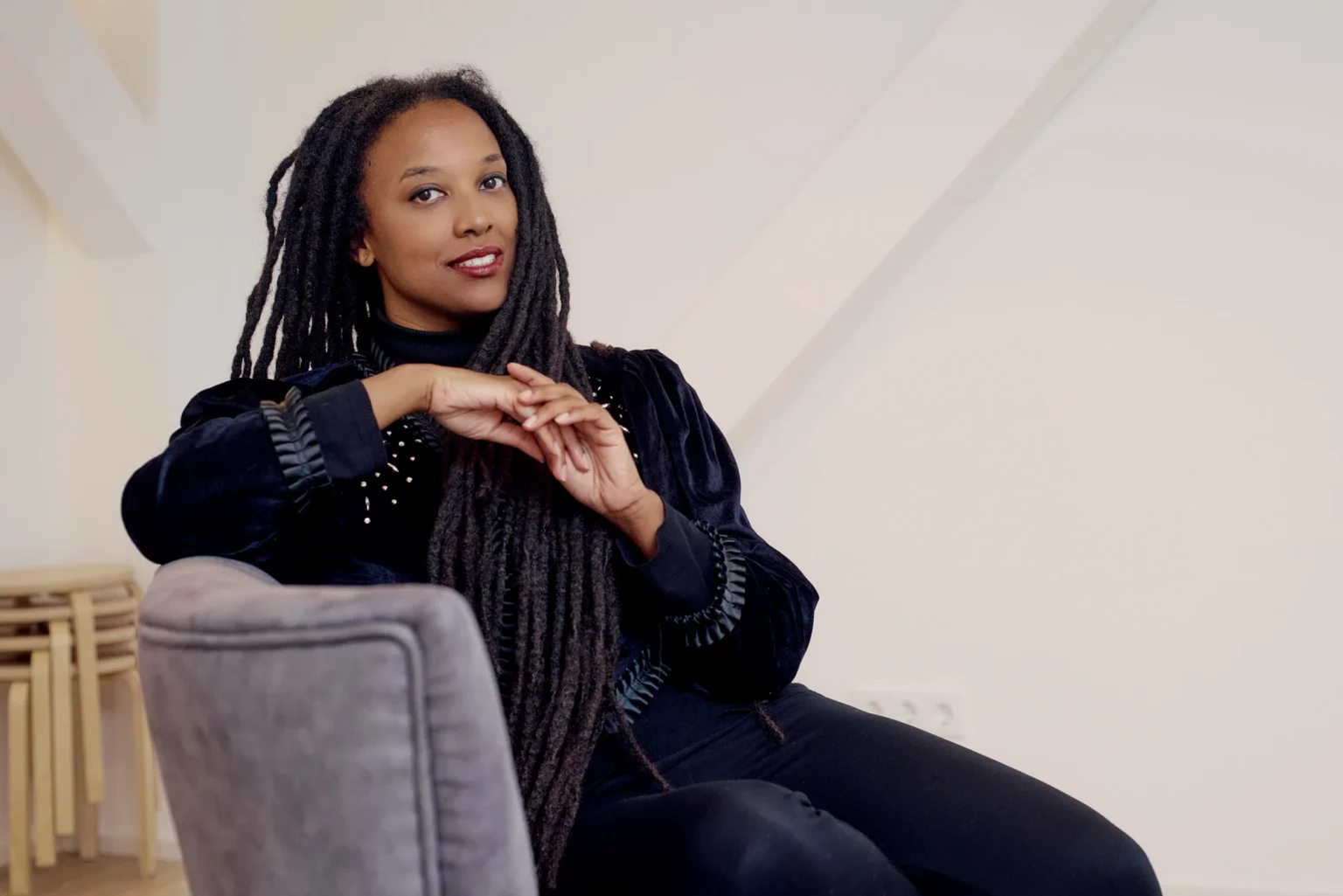 Eine Frau sitzt entspannt auf einem Stuhl und lehnt sich leicht zur Seite. Sie hat lange, dunkle Dreadlocks und trägt ein schwarzes Outfit. Ihre Hände sind locker ineinander verschränkt, und sie schaut mit einem ruhigen, freundlichen Ausdruck in die Kamera. Der Hintergrund ist hell und schlicht, mit ein paar gestapelten Hockern im Raum.