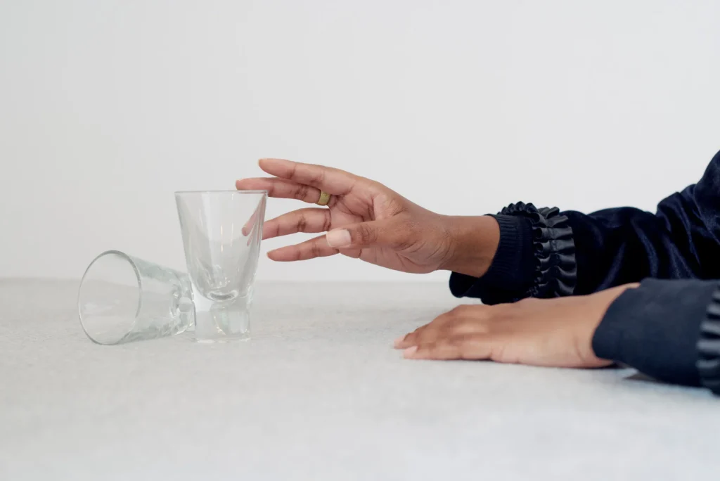 Eine Hand greift nach einem kleinen, durchsichtigen Glas, das auf einem Tisch steht. Daneben liegt ein weiteres Glas seitlich auf der Oberfläche. Die Szene ist schlicht gehalten, mit hellem Hintergrund und Fokus auf die Hände und die Gläser.