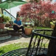 Mann sitzt entspannt im sonnigen Garten vor einem Wohnhaus, umgeben von Pflanzen, Bäumen und ruhiger Atmosphäre.