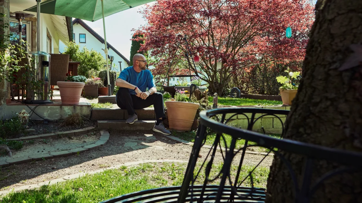 Mann sitzt entspannt im sonnigen Garten vor einem Wohnhaus, umgeben von Pflanzen, Bäumen und ruhiger Atmosphäre.