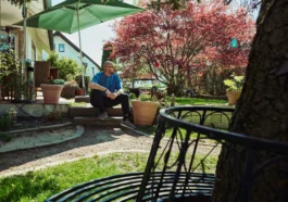Mann sitzt entspannt im sonnigen Garten vor einem Wohnhaus, umgeben von Pflanzen, Bäumen und ruhiger Atmosphäre.