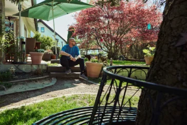 Mann sitzt entspannt im sonnigen Garten vor einem Wohnhaus, umgeben von Pflanzen, Bäumen und ruhiger Atmosphäre.