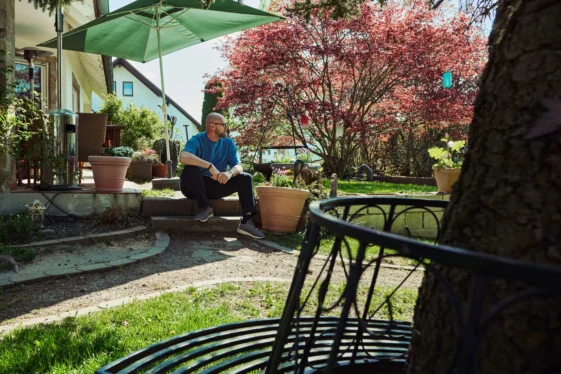 Mann sitzt entspannt im sonnigen Garten vor einem Wohnhaus, umgeben von Pflanzen, Bäumen und ruhiger Atmosphäre.