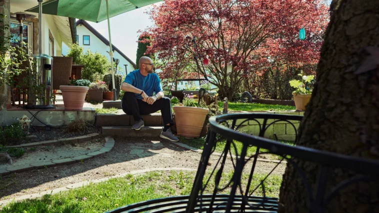 Mann sitzt entspannt im sonnigen Garten vor einem Wohnhaus, umgeben von Pflanzen, Bäumen und ruhiger Atmosphäre.