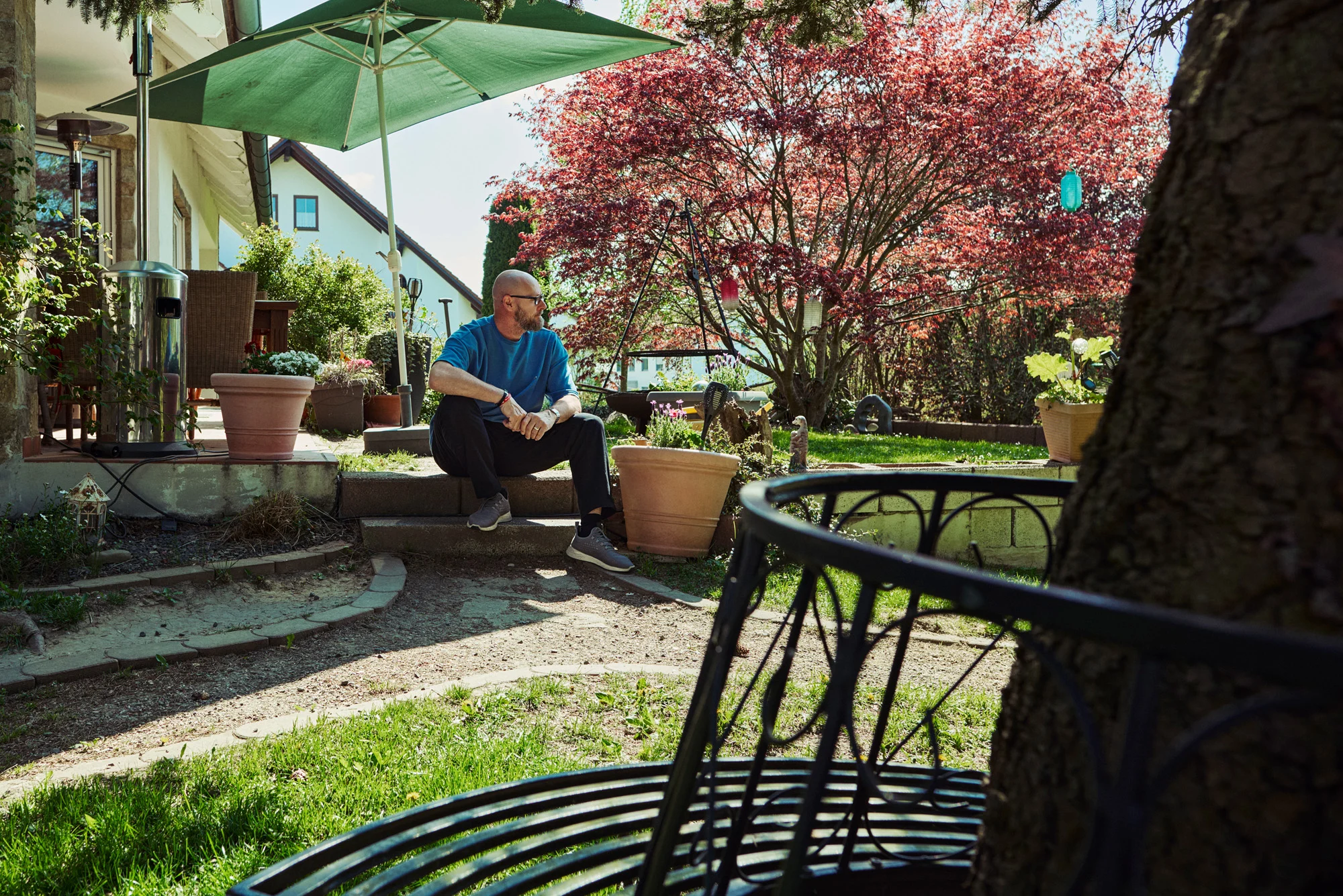 Mann sitzt entspannt im sonnigen Garten vor einem Wohnhaus, umgeben von Pflanzen, Bäumen und ruhiger Atmosphäre.