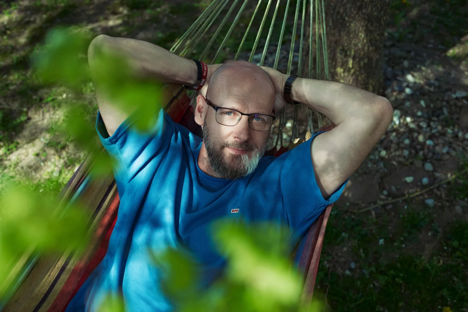 Mann mit Brille entspannt in einer Hängematte im Grünen, aufgenommen im natürlichen Sonnenlicht zwischen unscharfen Blättern.
