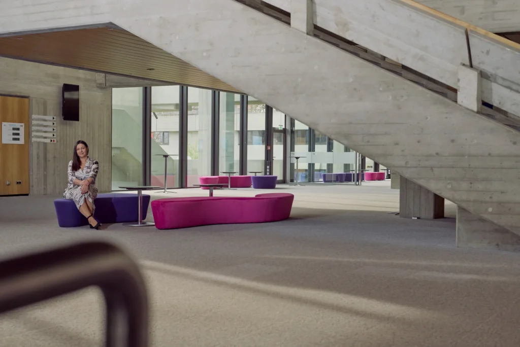 Frau sitzt entspannt auf einer violett-pinken Sitzbank in einem modernen, lichtdurchfluteten Gebäude mit Betonarchitektur und großer Treppe im Hintergrund.