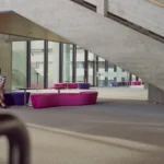 Frau sitzt entspannt auf einer violett-pinken Sitzbank in einem modernen, lichtdurchfluteten Gebäude mit Betonarchitektur und großer Treppe im Hintergrund.