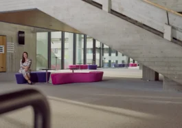 Frau sitzt entspannt auf einer violett-pinken Sitzbank in einem modernen, lichtdurchfluteten Gebäude mit Betonarchitektur und großer Treppe im Hintergrund.