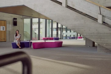 Frau sitzt entspannt auf einer violett-pinken Sitzbank in einem modernen, lichtdurchfluteten Gebäude mit Betonarchitektur und großer Treppe im Hintergrund.