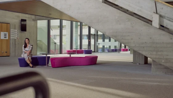 Frau sitzt entspannt auf einer violett-pinken Sitzbank in einem modernen, lichtdurchfluteten Gebäude mit Betonarchitektur und großer Treppe im Hintergrund.