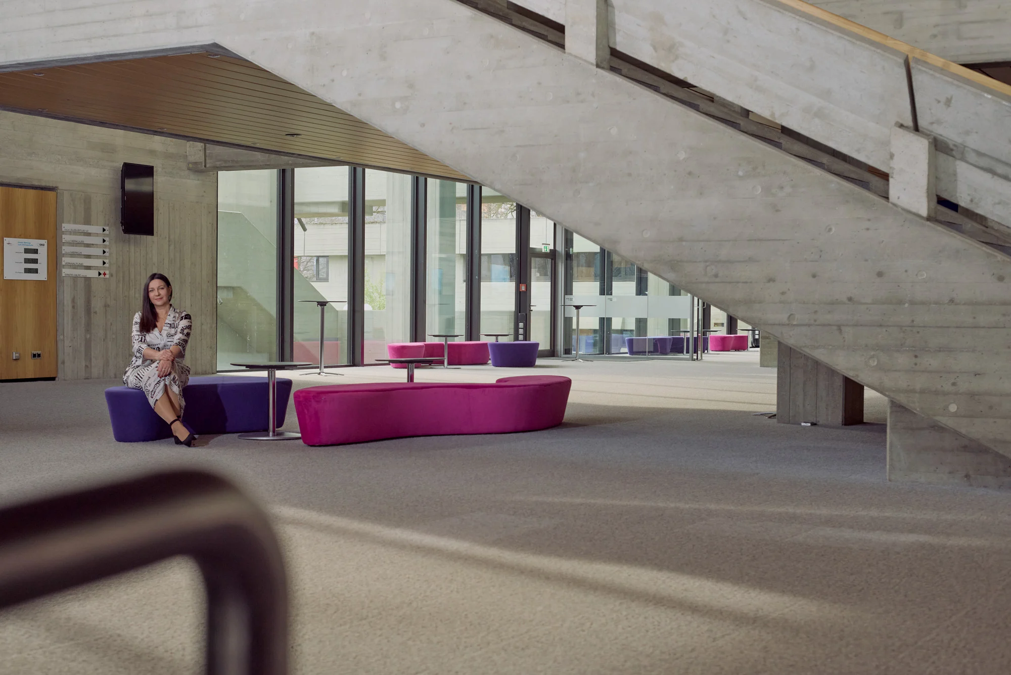 Frau sitzt entspannt auf einer violett-pinken Sitzbank in einem modernen, lichtdurchfluteten Gebäude mit Betonarchitektur und großer Treppe im Hintergrund.