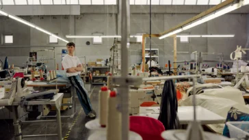 Porträt eines jungen Mannes in den Produktionshallen eines nachhaltigen Textilunternehmens, umgeben von Nähmaschinen, Stoffen und Arbeitsplätzen.
