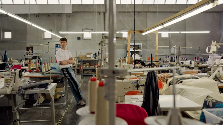 Porträt eines jungen Mannes in den Produktionshallen eines nachhaltigen Textilunternehmens, umgeben von Nähmaschinen, Stoffen und Arbeitsplätzen.