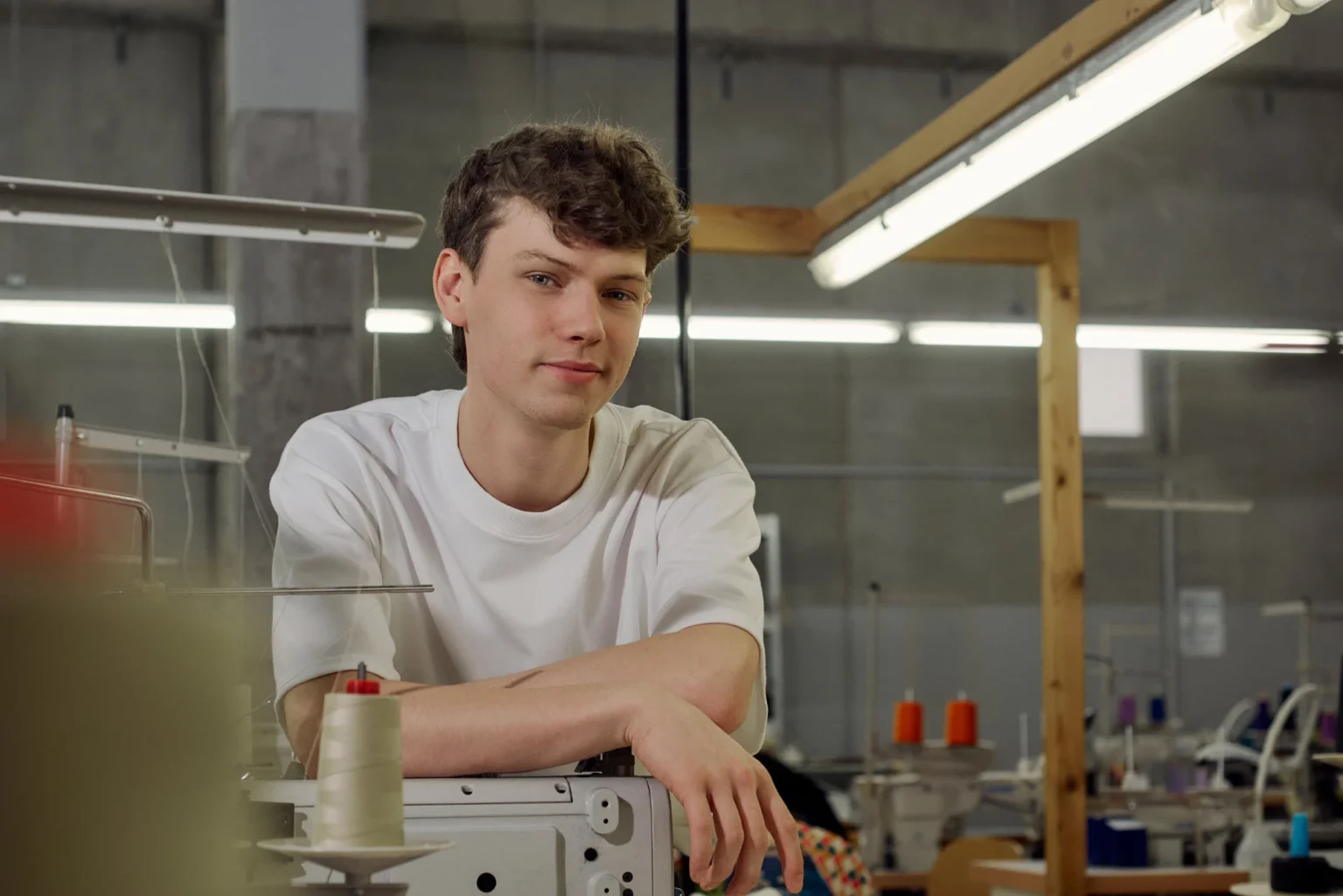 Porträt eines jungen Mannes in den Produktionshallen eines nachhaltigen Textilunternehmens, umgeben von Nähmaschinen, Stoffen und Arbeitsplätzen. Er blick leicht Frech in die Kamera und macht einen selbstbewussten Eindruck.