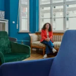 Porträt einer Frau im roten Blazer in einem stilvoll eingerichteten Innenraum mit farbenfrohen Sesseln, großen Fenstern und entspannter Atmosphäre.