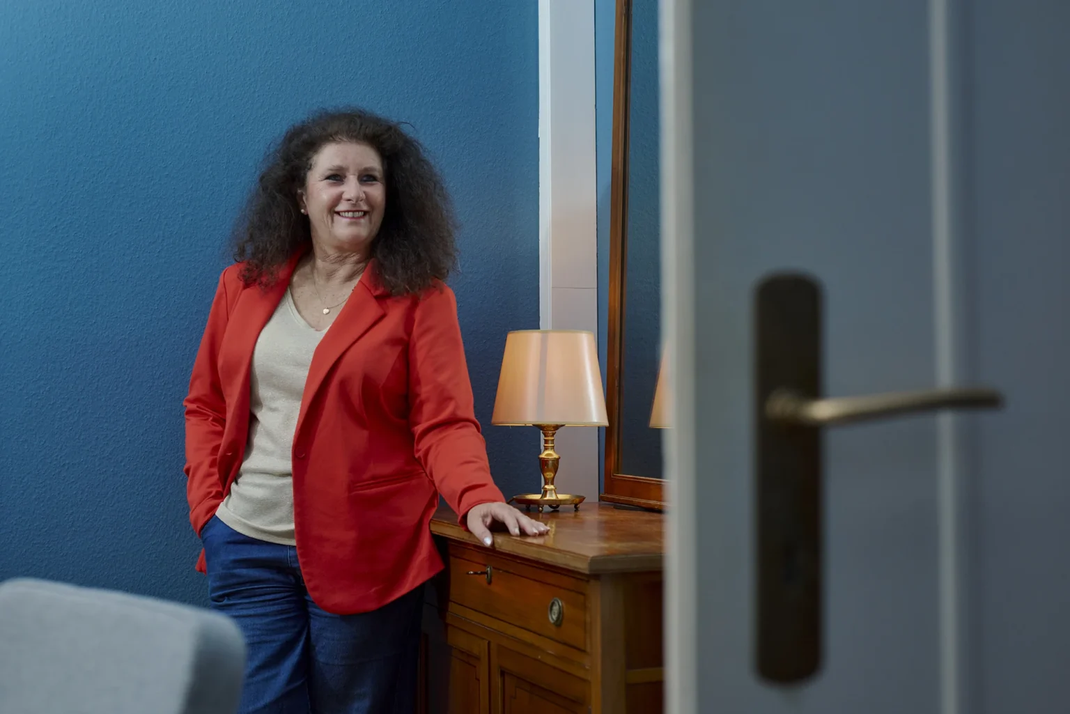 Porträt einer Frau im roten Blazer angelehnt an eine Kommode aus Holz in einem stilvoll eingerichteten Innenraum mit farbenfrohen Sesseln, großen Fenstern und entspannter Atmosphäre. Ihr Blick ist gelöst, energetisch und zur Kamera gerichtet.