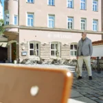 Businessporträt eines Mannes vor dem Restaurant Emmi’s Kitchen in Augsburg. Er steht auf einem historischen Platz mit Blick zur Seite, eingerahmt von Altstadtarchitektur und einer Kirche im Hintergrund.