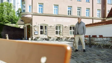 Businessporträt eines Mannes vor dem Restaurant Emmi’s Kitchen in Augsburg. Er steht auf einem historischen Platz mit Blick zur Seite, eingerahmt von Altstadtarchitektur und einer Kirche im Hintergrund.