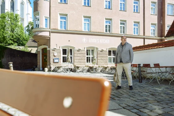 Businessporträt eines Mannes vor dem Restaurant Emmi’s Kitchen in Augsburg. Er steht auf einem historischen Platz mit Blick zur Seite, eingerahmt von Altstadtarchitektur und einer Kirche im Hintergrund.