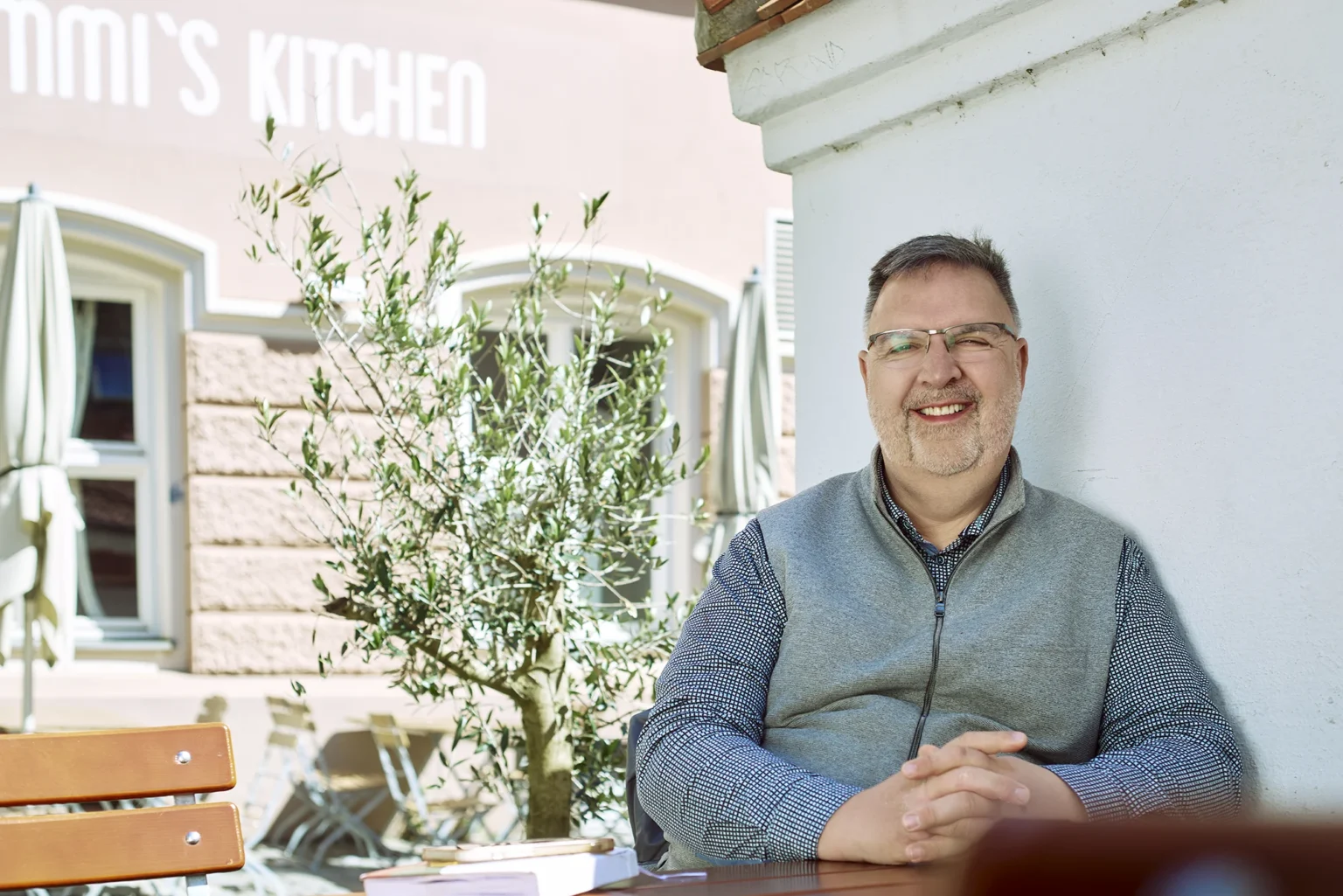 Andreas Becker sitzt lächelnd an einem Tisch in der Augsburger Altstadt. Der Unternehmensberater trägt ein graues Fleece-Gilet über einem gemusterten Hemd und lehnt entspannt an einer hellen Mauer. Im Hintergrund sind ein Olivenbaum, Café-Mobiliar und die Fassade eines Restaurants zu sehen, wodurch eine freundliche und entspannte Atmosphäre entsteht.