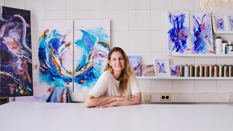 Eine Künstlerin sitzt in ihrem hellen Atelier an einem weißen Tisch. Hinter ihr hängen zahlreiche farbenfrohe abstrakte Gemälde in Blau, Türkis, Violett und Gold. Auf einem Regal stehen weitere Bilder und Farbspraydosen. Die Künstlerin blickt direkt in die Kamera.
